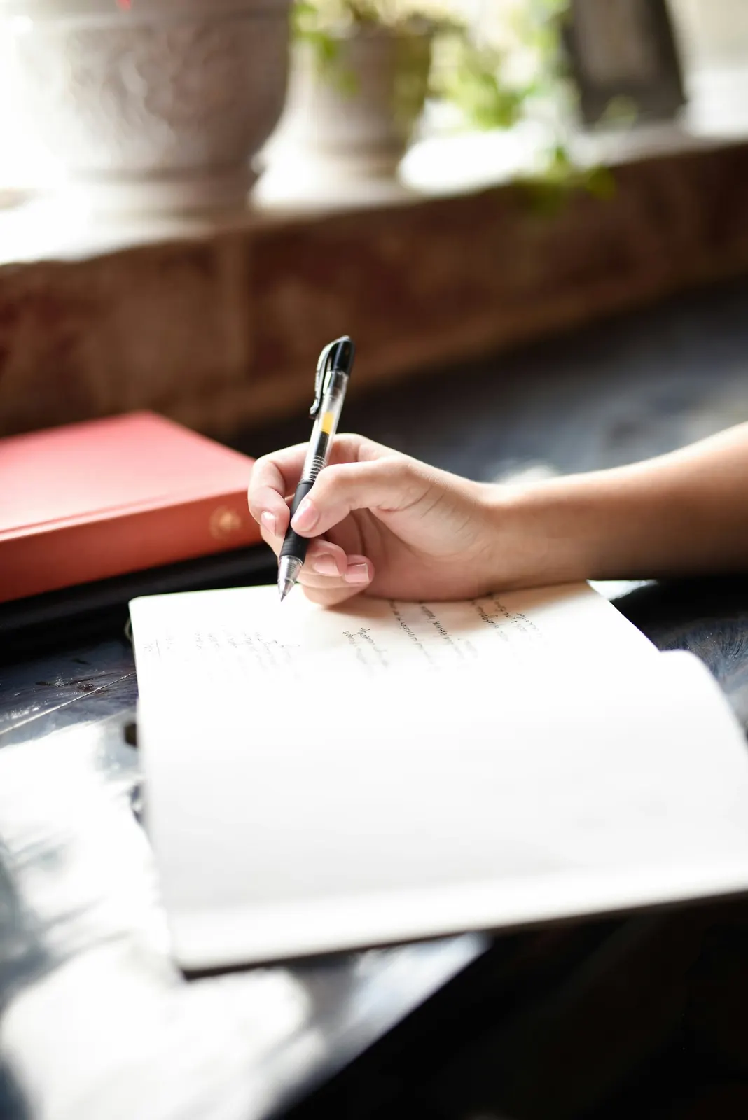 Une main écrit au stylo dans un carnet ouvert, près d'une fenêtre
